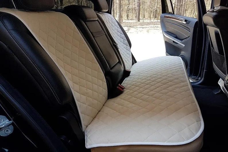 May include: Beige quilted car seat covers. The covers are diamond-stitched and designed to fit the back seats of a vehicle. The seats are installed in a black interior, with a door open in the background.