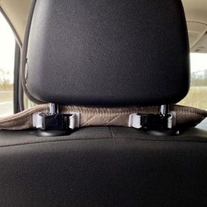 May include: A car seat headrest with a tan quilted cover and black plastic clips. The headrest is black and the seat is a dark gray fabric. The cover is secured with white elastic straps.