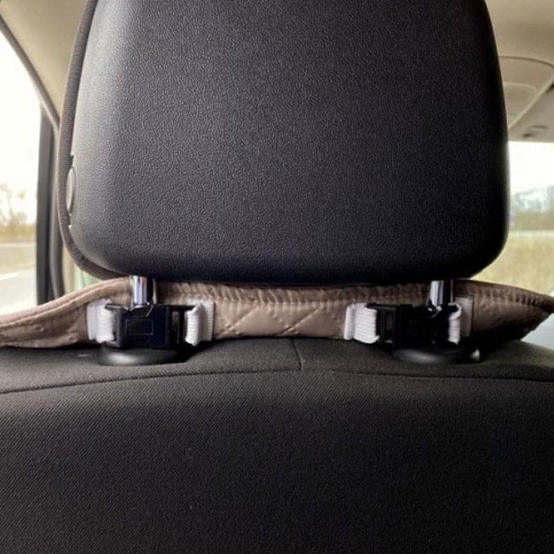 May include: A car seat headrest with a tan quilted cover and black plastic clips. The headrest is black and the seat is a dark gray fabric. The cover is secured with white elastic straps.