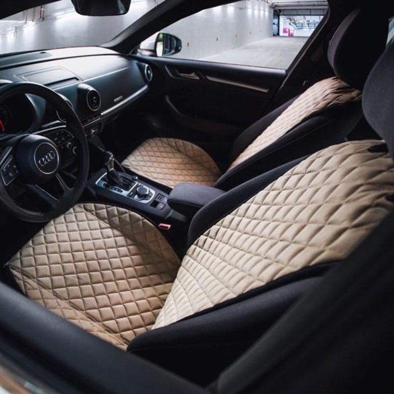 May include: Beige quilted car seat covers installed in a black interior. The covers have a diamond pattern and are visible on the front and back seats. The car's steering wheel and dashboard are also visible.