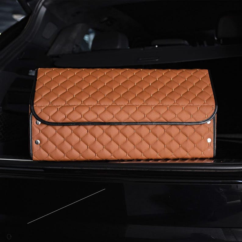 May include: A brown quilted car trunk organizer with black trim and silver snaps. The organizer is folded and sitting in the back of a car.
