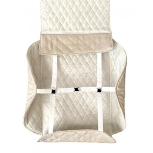May include: Cream-colored quilted car seat cover with diamond stitching. Features a tan border and white straps with black buckles for secure attachment. The cover is designed to protect the car seat.