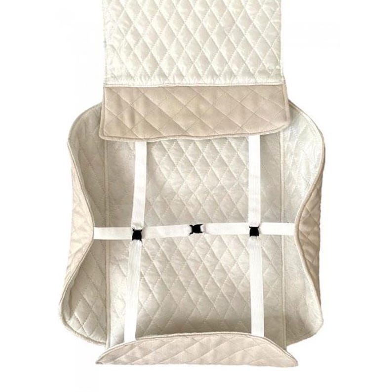 May include: Cream-colored quilted car seat cover with diamond stitching. Features a tan border and white straps with black buckles for secure attachment. The cover is designed to protect the car seat.