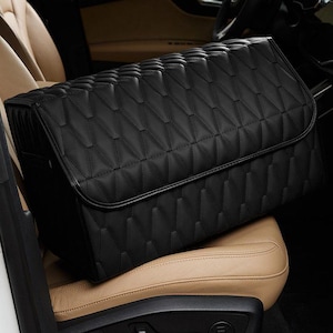 K&ouml;nnte beinhalten: Ein schwarzer, gesteppter Auto-Organizer mit geometrischem Muster, der auf einem beigefarbenen Autositz liegt. Der Organizer hat einen Klappenverschluss und besteht aus einem glatten, lederartigen Material. Der Innenraum eines Autos ist im Hintergrund sichtbar.