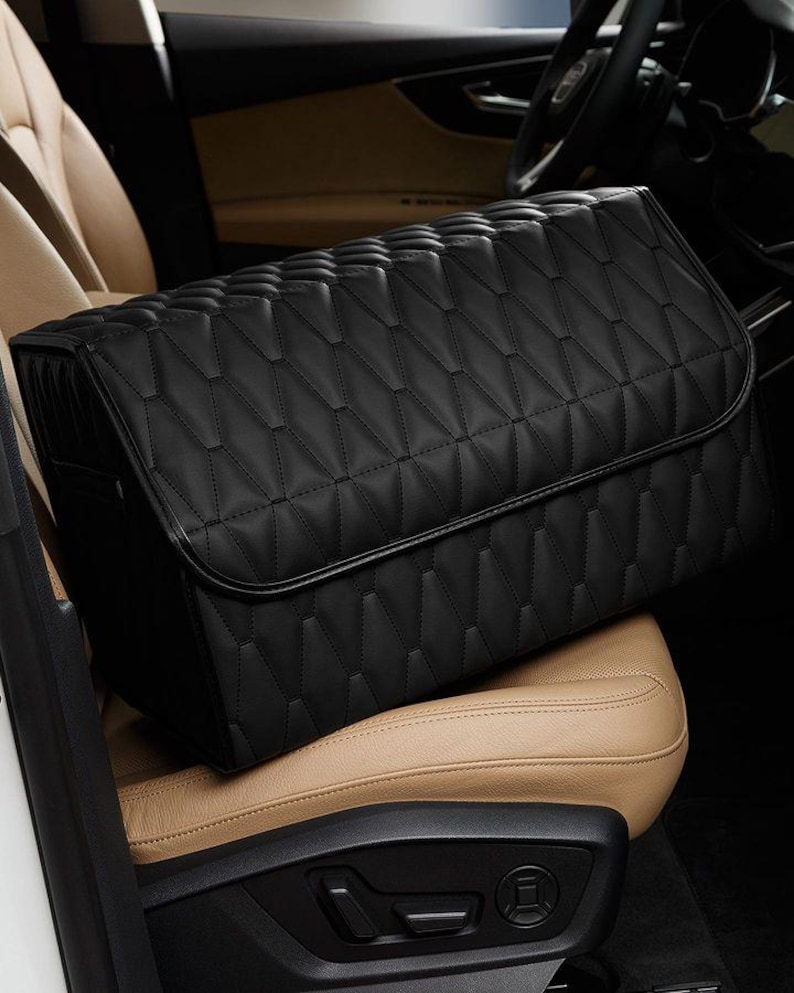 K&ouml;nnte beinhalten: Ein schwarzer, gesteppter Auto-Organizer mit geometrischem Muster, der auf einem beigefarbenen Autositz liegt. Der Organizer hat einen Klappenverschluss und besteht aus einem glatten, lederartigen Material. Der Innenraum eines Autos ist im Hintergrund sichtbar.