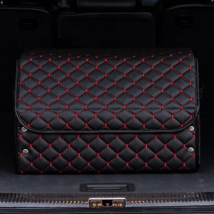 May include: Black quilted storage box with red stitching. The box is in the trunk of a car.