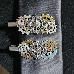Puede incluir: Dos pinzas de corbata de estilo steampunk con diseños de rosa de los vientos y engranajes. Las pinzas presentan engranajes plateados, bronce y dorados, y una rosa de los vientos en el centro. Las pinzas se muestran sobre una corbata gris oscuro.