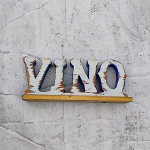 May include: White wooden sign with the word "VINO" painted in blue letters. The sign is on a white background.