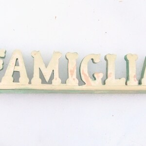 May include: A wooden sign with the word "FAMIGLIA" painted in white with green outlining. The sign is on a white background.