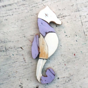 May include: A wooden seahorse ornament painted in white, purple, and yellow. The seahorse is standing on a white wooden surface.