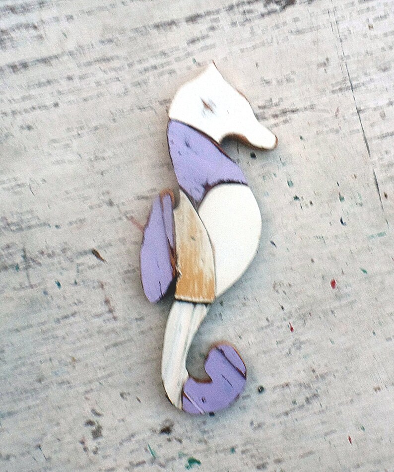 May include: A wooden seahorse ornament painted in white, purple, and yellow. The seahorse is standing on a white wooden surface.