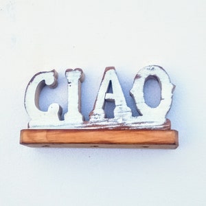 May include: Wooden sign with the word "CIAO" painted in white on a distressed wood background. The sign is mounted on a wooden base.