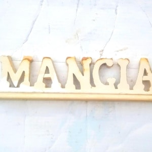 May include: A wooden sign with the word "MANGIA" carved into it. The sign is painted white and has a natural wood finish.
