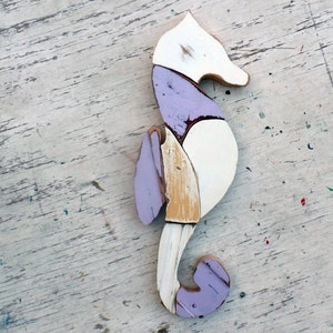 May include: A wooden seahorse sculpture painted in shades of white, purple, and brown. The seahorse is made of multiple pieces of wood that have been joined together to create a unique and whimsical design.