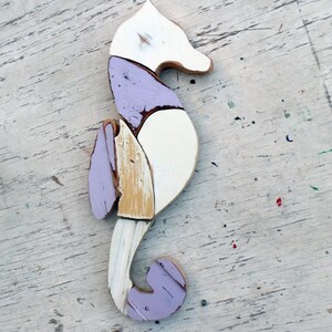 May include: A wooden seahorse sculpture painted in shades of white, purple, and yellow. The seahorse is made of multiple pieces of wood that have been joined together.