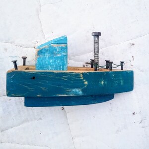 Pu&ograve; includere: Una piccola barca di legno dipinta di blu-verde con una cabina in legno blu e bianca. La barca ha una vite di metallo per l'albero maestro e diversi chiodi di metallo per una ringhiera.