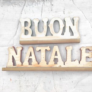 May include: Two wooden signs with the words "BUON NATALE" in a decorative font. The letters are painted white with a brown trim, and the signs are set against a textured, light-colored background.