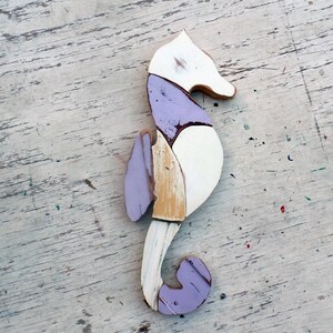 May include: A wooden seahorse sculpture painted in white, purple, and beige. The seahorse is made of multiple pieces of wood that have been joined together.