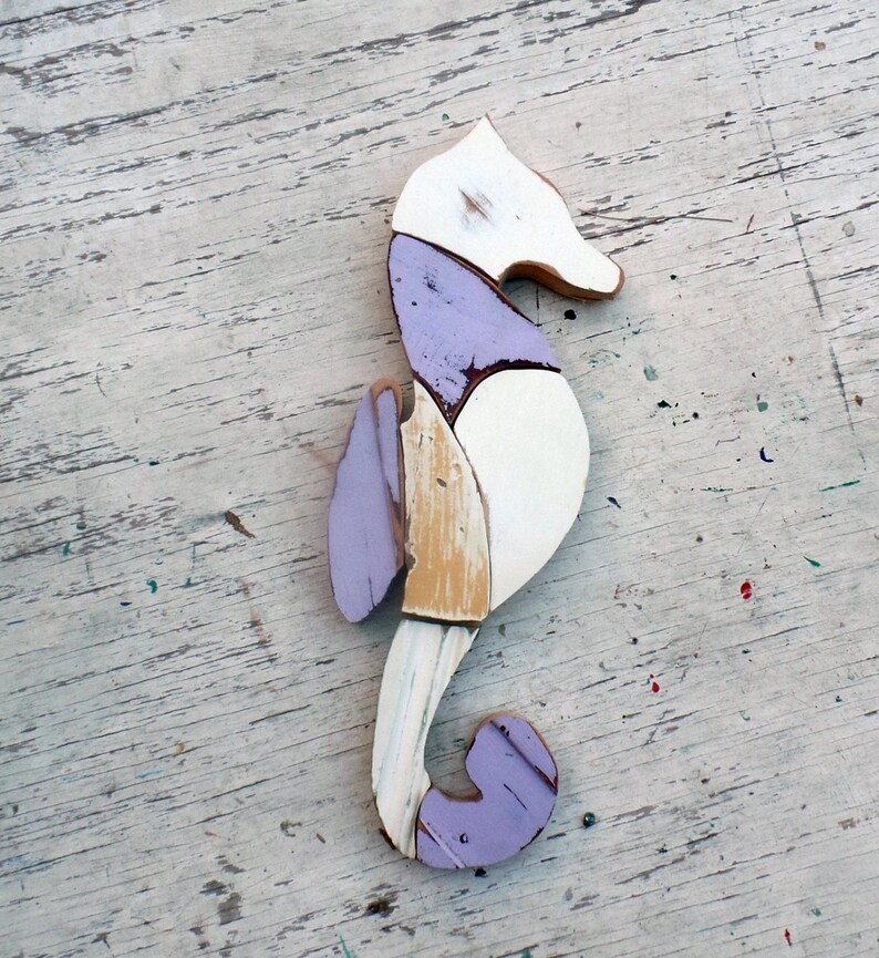 May include: A wooden seahorse sculpture painted in white, purple, and beige. The seahorse is made of multiple pieces of wood that have been joined together.