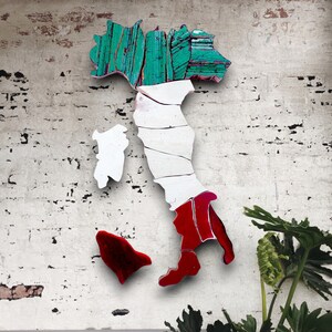 May include: A wooden cutout of the Italian peninsula painted in the colors of the Italian flag. The boot of Italy is red, the middle section is white, and the top section is green. The cutout is mounted on a white, textured wall.