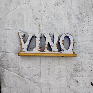 May include: White wooden sign with the word "VINO" painted in blue letters. The sign has a yellow wooden base.