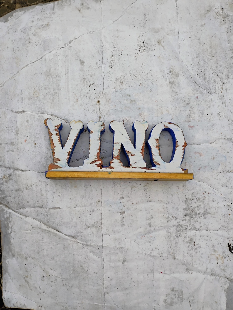May include: White wooden sign with the word "VINO" painted in blue letters. The sign has a yellow wooden base.