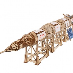 May include: A wooden model of the Saturn V rocket, a multi-stage rocket used in the Apollo programme. The model is detailed and includes the iconic "USA" markings. It is displayed on a wooden stand.