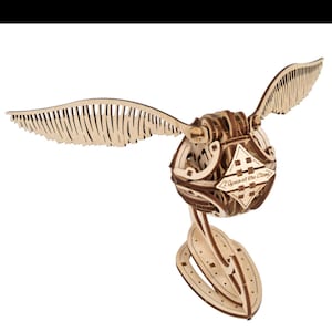 Könnte beinhalten: Ein hölzernes Modell des Goldenen Schnatz mit detaillierten Flügeln und einem kugelförmigen Körper. Die Flügel sind aufwendig mit parallelen Linien geschnitten. Der Körper hat ein rautenförmiges Emblem mit dem Text "I Open at the Close".