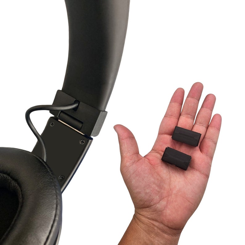 May include: Black over-ear headphones with a black headband and earcups. Two small black rectangular foam pads are held in a hand. The headphones have a cable attached to the left earcup.