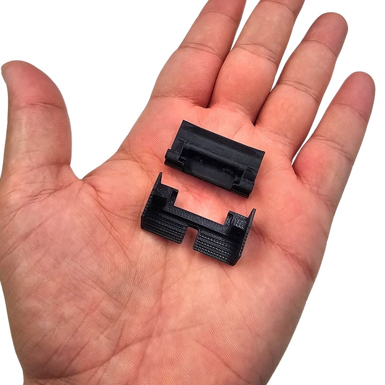 May include: Two small, black plastic components are displayed. The parts have a rectangular shape with a grooved texture on the bottom. One piece is positioned above the other, suggesting they may connect or fit together.