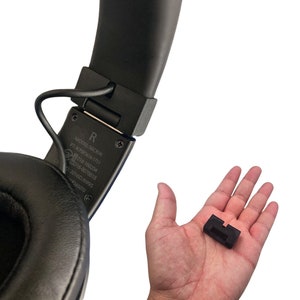 May include: Black over-ear headphones with a small, rectangular black accessory held in a hand. The headphones have a padded ear cup and a black headband. The accessory appears to be a connector or adapter.
