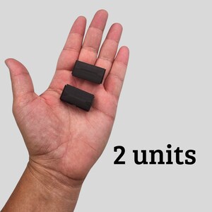 May include: Two small, dark rectangular objects are held in an open hand against a light gray background. The text "2 units" is displayed in black, bold font below the hand.