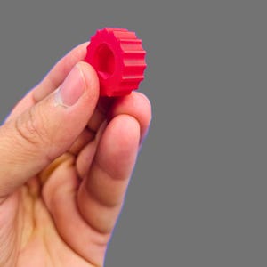 May include: A bright red plastic gear with a hole in the center. The gear has a textured surface and is held in a person's hand.