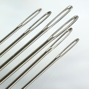 May include: Five silver sewing needles with eye holes at the top. The needles are arranged in a fan shape, with the largest needle at the top and the smallest needle at the bottom.