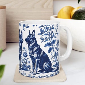 May include: White ceramic mug with a blue and white design featuring two German Shepherd dogs and floral patterns. The mug has a white handle.