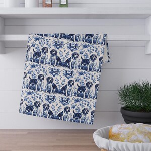 May include: A white and blue patterned kitchen towel featuring a repeating design of dogs and floral elements. The dogs are depicted in a seated position. The towel is hanging on a white shelf above a wooden countertop.