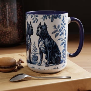 May include: White ceramic mug with a blue interior and handle, featuring a blue and white illustration of two dogs and floral patterns. The mug is on a wooden surface with a spoon and a pastry.