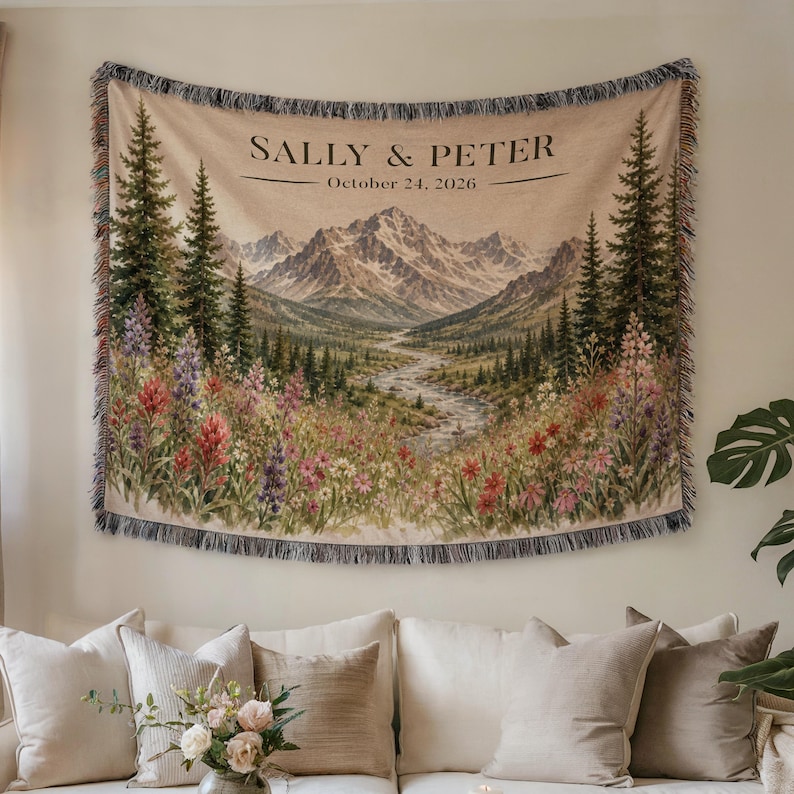 May include: A woven wall tapestry featuring a mountain landscape with pine trees, a winding river, and wildflowers. The text SALLY & PETER and October 24, 2026 is printed at the top. The tapestry has a fringed edge and a beige background.