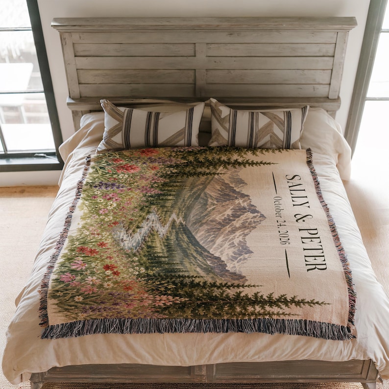 May include: A woven throw blanket featuring a landscape design with a mountain range, pine trees, and a field of colorful wildflowers. The text "SALLY & PETER" and "October 24, 2026" is printed on the right side. The blanket has a fringed edge.