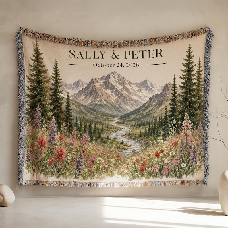 May include: A woven throw blanket featuring a mountain landscape with a winding river, pine trees, and colorful wildflowers. The text SALLY & PETER and October 24, 2026 is printed at the top. The blanket has a fringed edge with multi-colored threads.