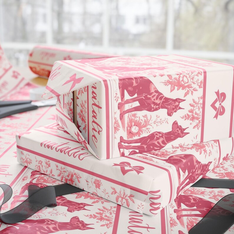 May include: Gift boxes wrapped in white paper with a pink floral and dog pattern. The boxes are stacked, with one partially unwrapped, revealing the word "natalie" in script. Black ribbon and scissors are also visible.