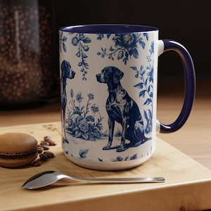 May include: White ceramic mug with a blue floral and dog design. The mug features a blue handle and rim. A dog illustration is centered on the mug, surrounded by blue floral patterns. The mug is on a wooden surface.