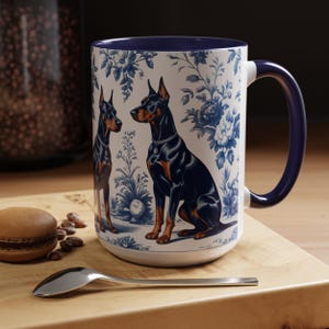 May include: A white ceramic mug with a blue rim and handle. The mug features a blue and white floral pattern with two black Doberman Pinschers.