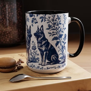 May include: A white ceramic mug with a black rim and handle. The mug features a blue and white floral pattern with two German Shepherd dogs. The dogs are sitting and looking at each other.
