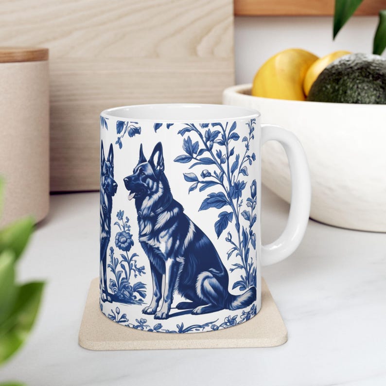 May include: White ceramic mug with a blue and white design featuring two German Shepherd dogs and floral patterns. The mug has a white handle.