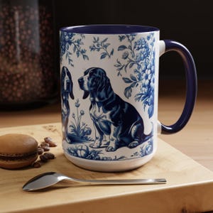 May include: White ceramic mug with a blue and white basset hound design. The mug features a blue handle and rim, and is decorated with floral patterns. A spoon and a pastry are on a wooden surface.