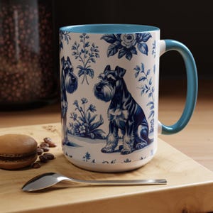 May include: A white ceramic mug with a blue interior and handle. The mug features a blue and white toile design with illustrations of Schnauzer dogs and floral elements. A spoon and a macaron are on a wooden surface.