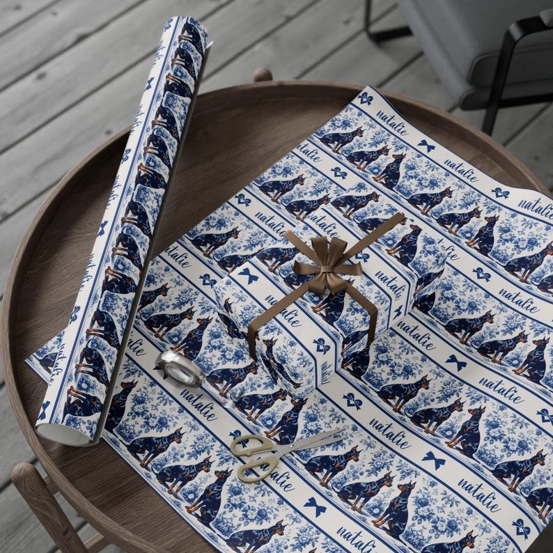 May include: Gift wrap featuring a repeating pattern of Doberman Pinschers in navy blue on a white background with floral accents. The word "natalie" is printed on the wrap. A roll of wrap, wrapped gift, scissors, and tape are on a wooden table.