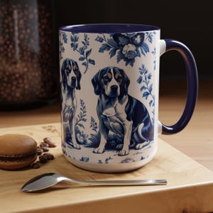 May include: A white ceramic mug with a blue and white floral pattern featuring beagles. The mug has a blue rim and handle.