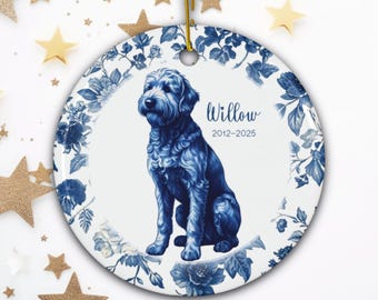 May include: A round, white ceramic ornament featuring a blue dog illustration. The dog is surrounded by a blue floral border. The ornament has the name "Willow" and the years "2012-2025" printed on it. A gold string is attached for hanging.